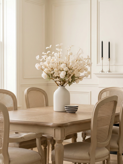 Dining room with wooden table and chairs, white vase with flowers, and candles.