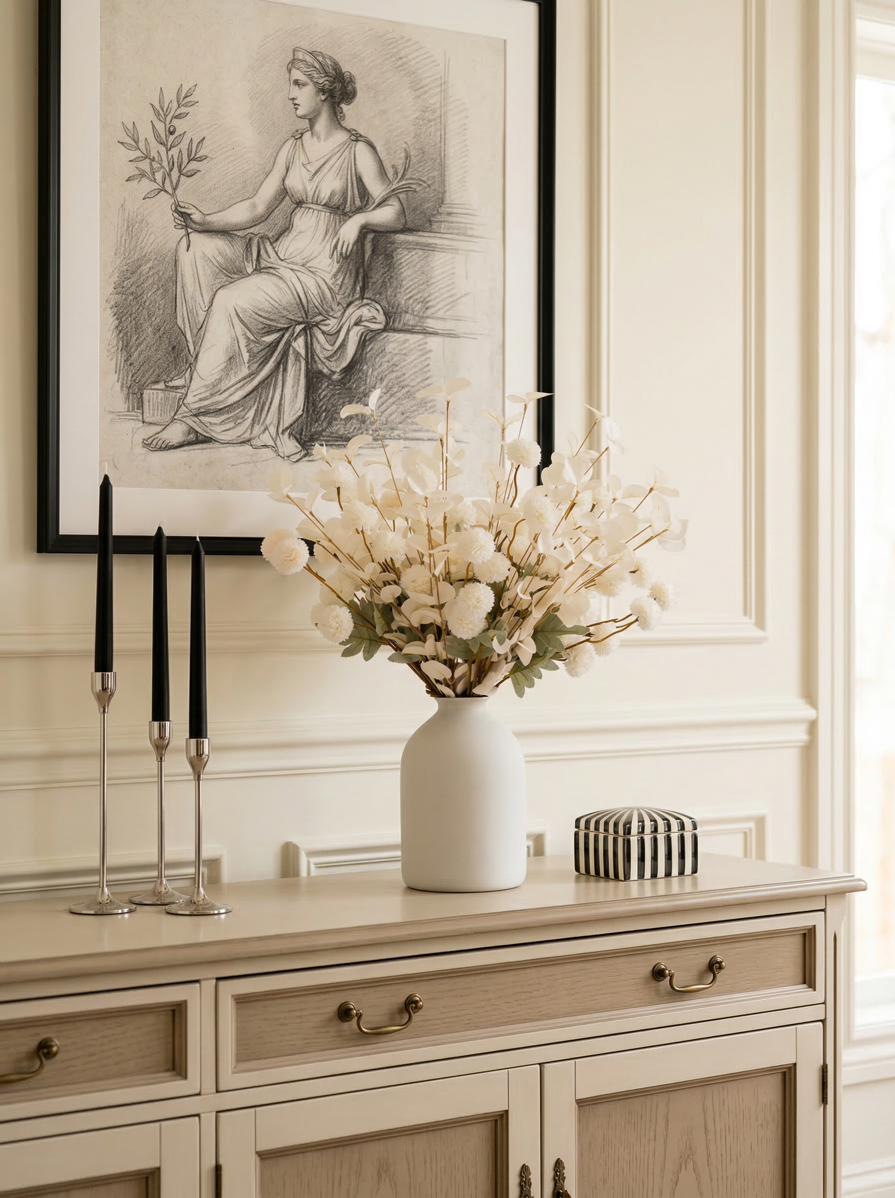 Decorative setting with a vase of flowers on a cabinet, framed artwork above, and black candlesticks.